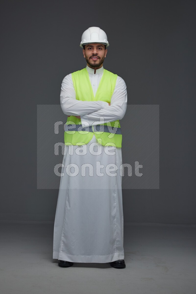 A Saudi man wearing Thobe with a yellow safety vest and white helmet standing and crossing his hands eye level on a gray background
