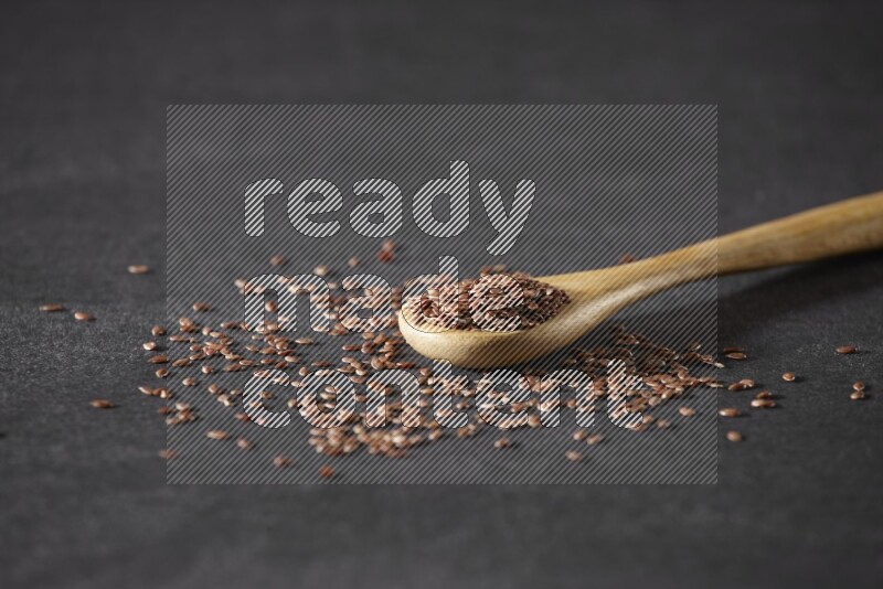 A wooden spoon full of flaxseeds and seeds spread beside it on a black flooring