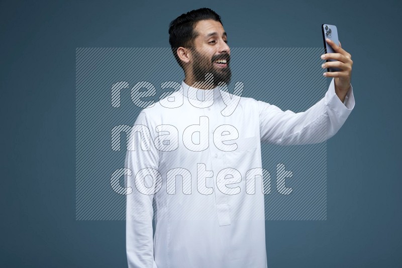A Saudi man Taking a Selfie in a blue background wearing Saudi Thob