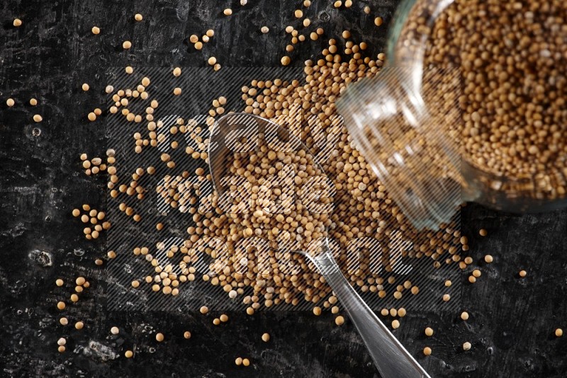 A glass spice jar and a metal spoon full of mustard seeds and jar is flipped with fallen seeds on a textured black flooring in different angles