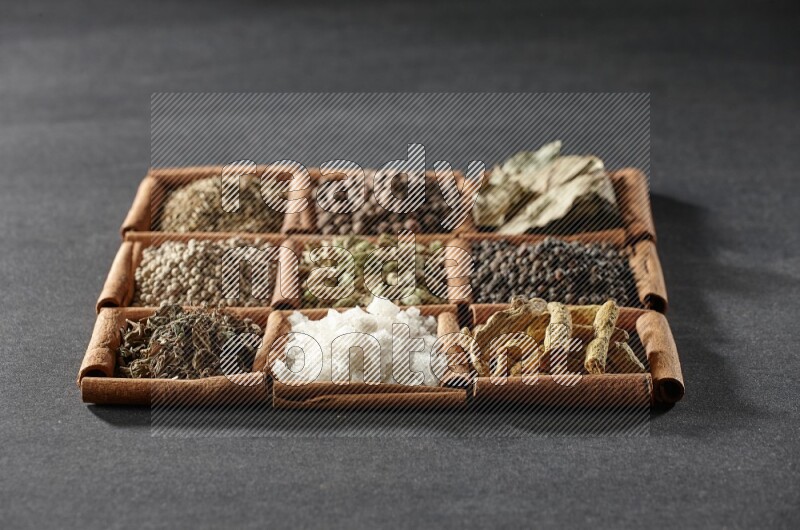 9 مربعات من أعواد القرفة ممتلئة بالهيل في المنتصف محاطة بأصابع الكركم والفلفل الأسود وورق الغار والبهار الحلو والكمون والفلفل الأبيض والريحان المجفف والملح على أرضية سوداء
