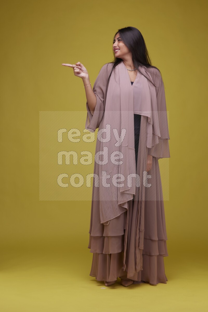 A woman Pointing on a Yellow Background wearing Brown Abaya