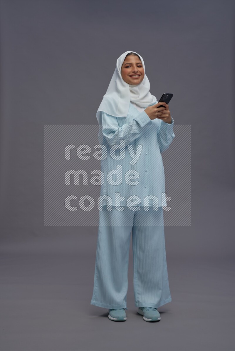 Saudi woman wearing hijab clothes standing texting on phone on gray background