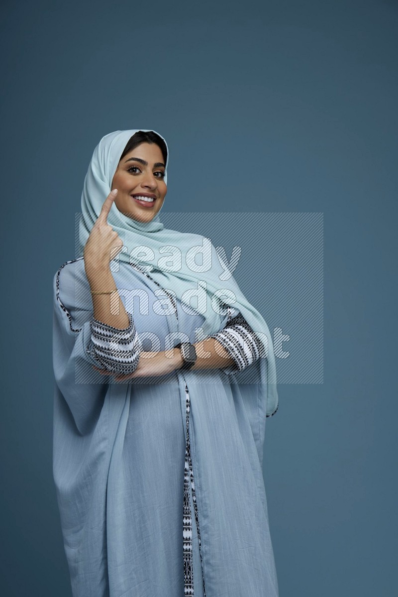 A Saudi woman pointing in a blue background wearing a blue Abaya with hijab