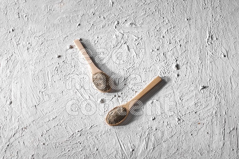 2 wooden spoons full of black pepper powder on a textured white background