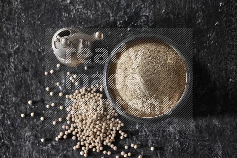 وعاء فخار أسود مملوء ببودرة الفلفل الأبيض مع حبات الفلفل ومطحنة الفلفل المعدنية على أرضية سوداء