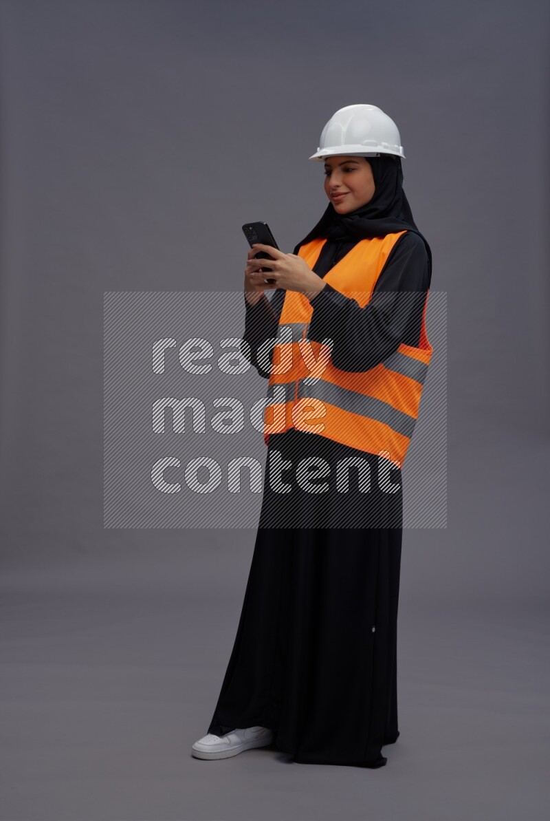 Saudi woman wearing Abaya with engineer vest standing texting on phone on gray background
