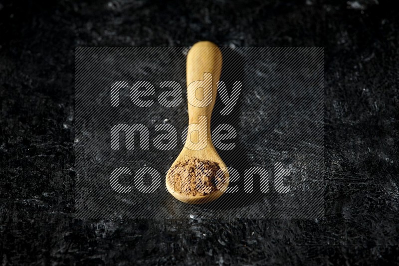 A wooden spoon full of allspice powder on a textured black flooring