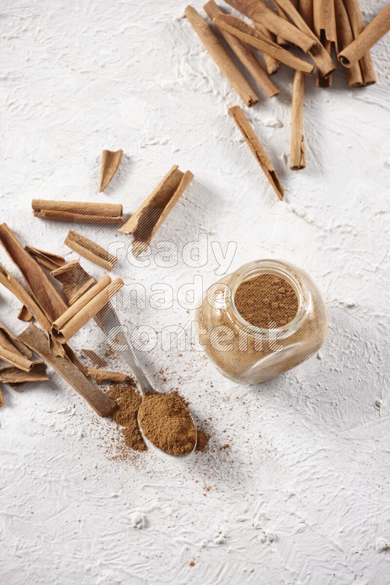 Herbal glass jar full cinnamon powder and a metal spoon surrounded by cinnamon sticks on a white background