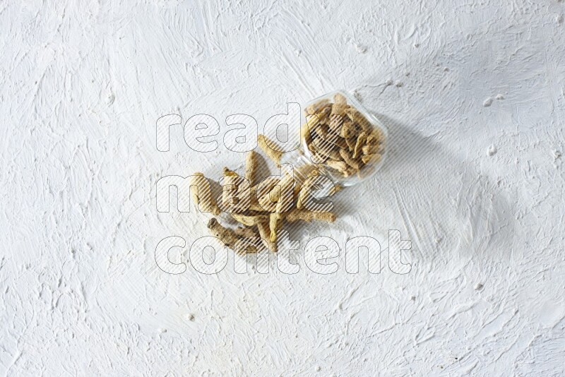 A flipped glass spice jar full of dried turmeric fingers and fell out it on textured white flooring