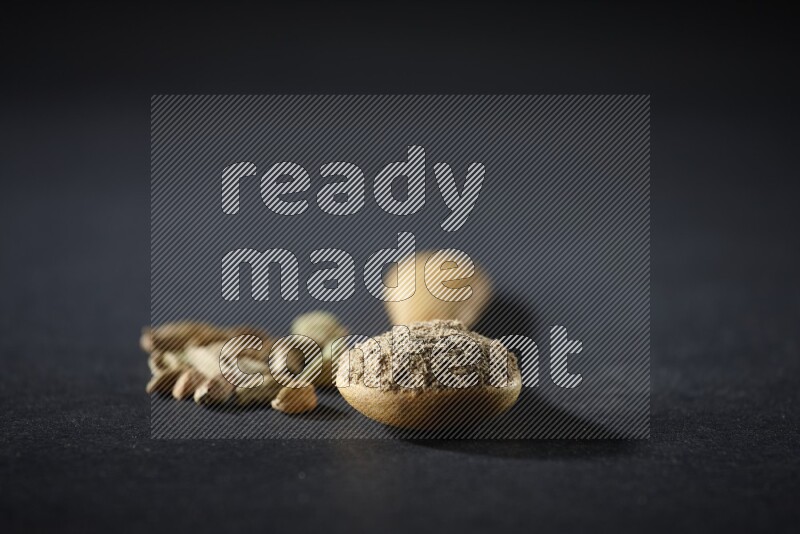 A wooden spoon full of cardamom powder and cardamom seeds beside it on black flooring