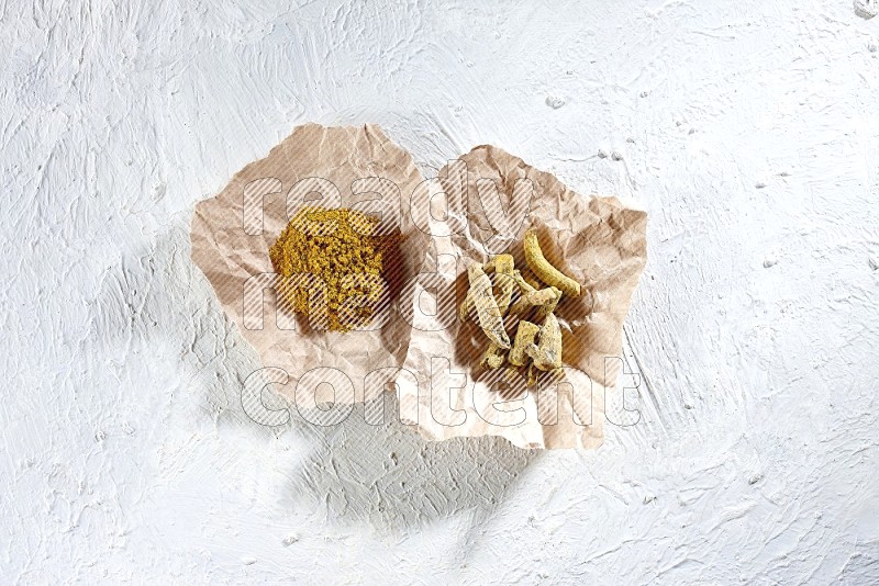 Turmeric powder and dried turmeric whole fingers in 2 crumpled pieces of paper on textured white flooring