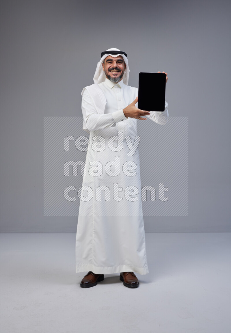 Saudi man Wearing Thob and white Shomag standing showing tablet to camera on Gray background