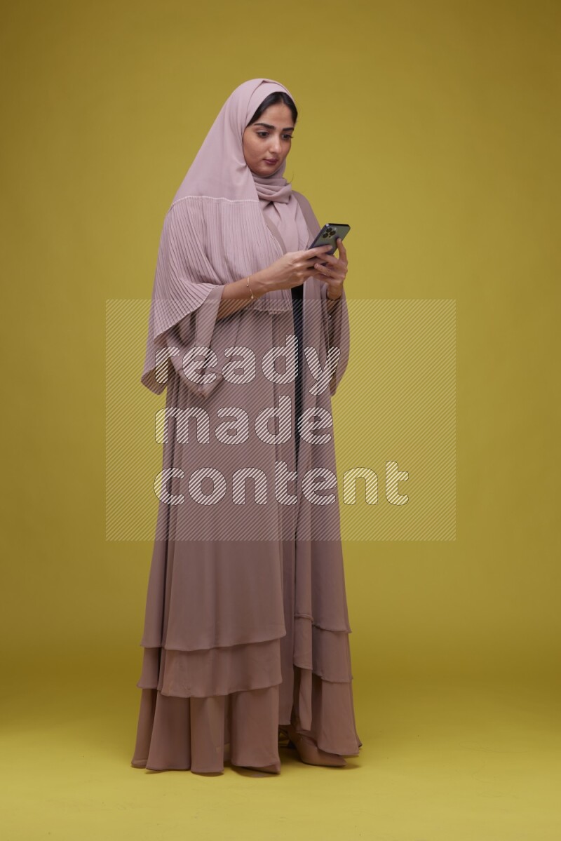 A woman Texting on her phone on a Yellow Background wearing Brown Abaya with Hijab