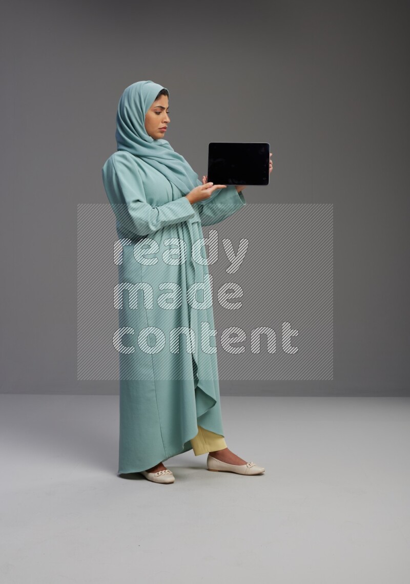 Saudi Woman wearing Abaya standing showing tablet to camera on Gray background