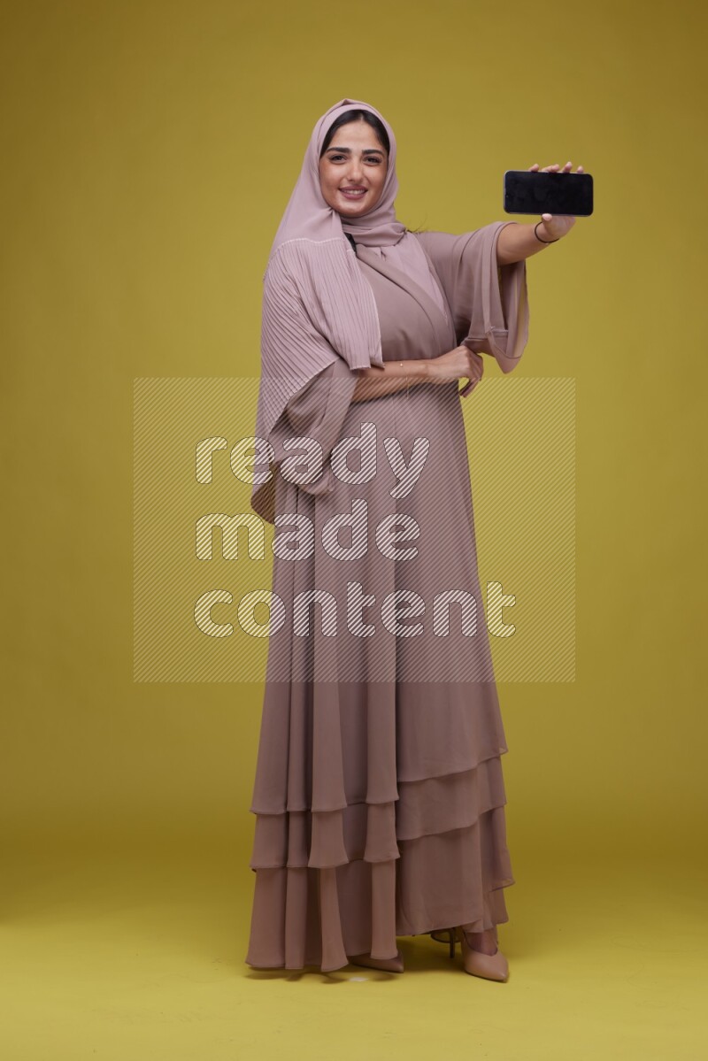 A woman Showing her Phone screen on a Yellow Background wearing Brown Abaya with Hijab