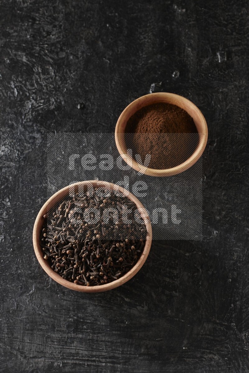 2 wooden bowls full of cloves powder and whole cloves on a textured black flooring