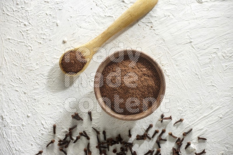 A wooden bowl and wooden spoon full of cloves powder with cloves spread on textured white flooring