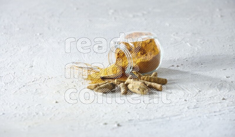 A flipped glass spice jar and metal spoon full of turmeric powder and powder spilled out of it with dried whole fingers on textured white flooring