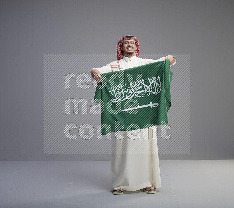 A saudi man standing wearing thob and red shomag holding big saudi flag on gray background