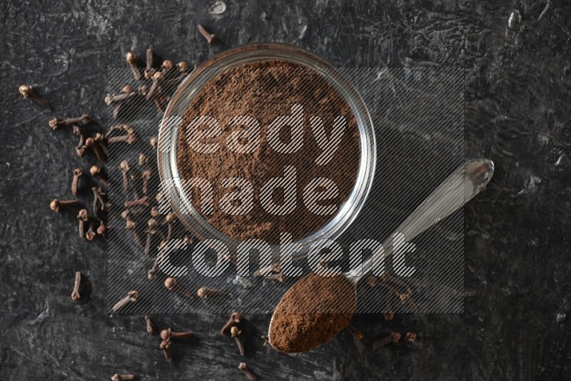 وعاء زجاجي وملعقة معدنية ممتلئان ببودرة القرنفل مع حبوب القرنفل بجانبهما على خلفية سوداء