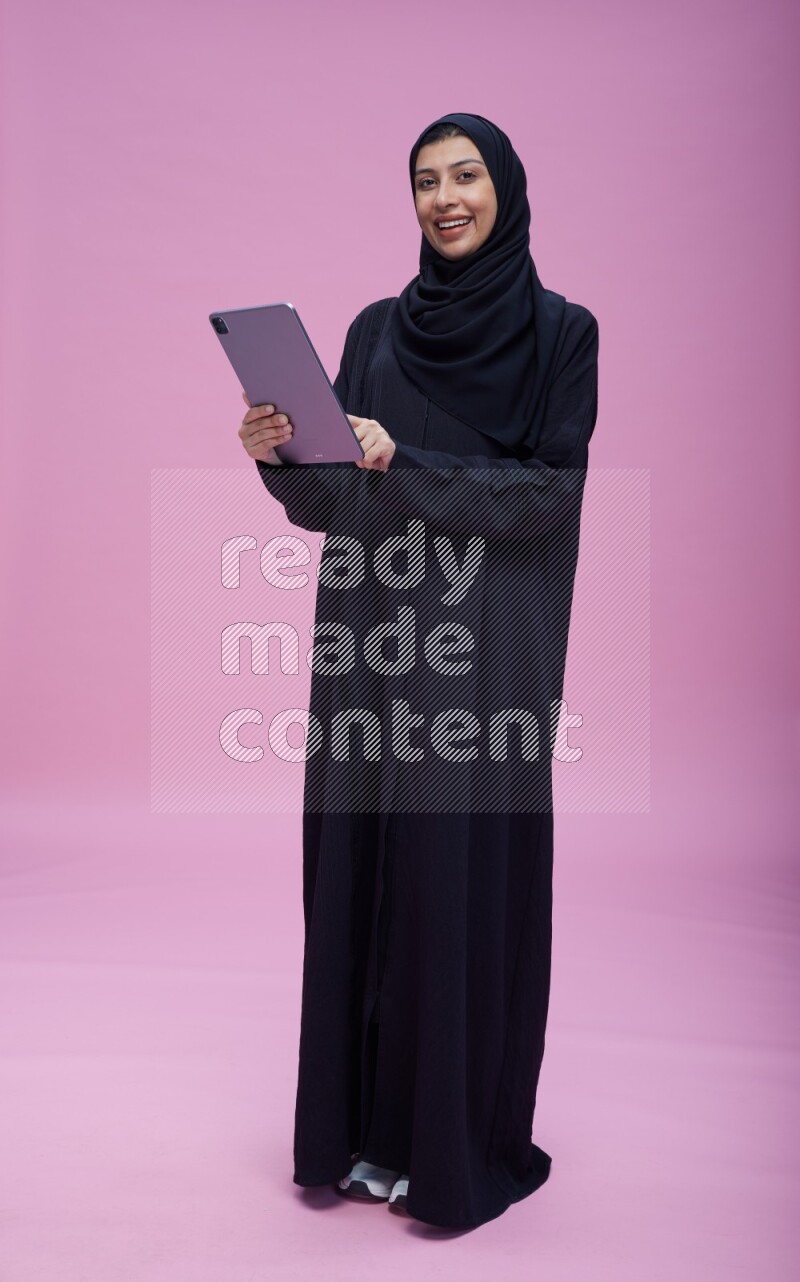 Saudi woman wearing Abaya standing working on tablet on pink background