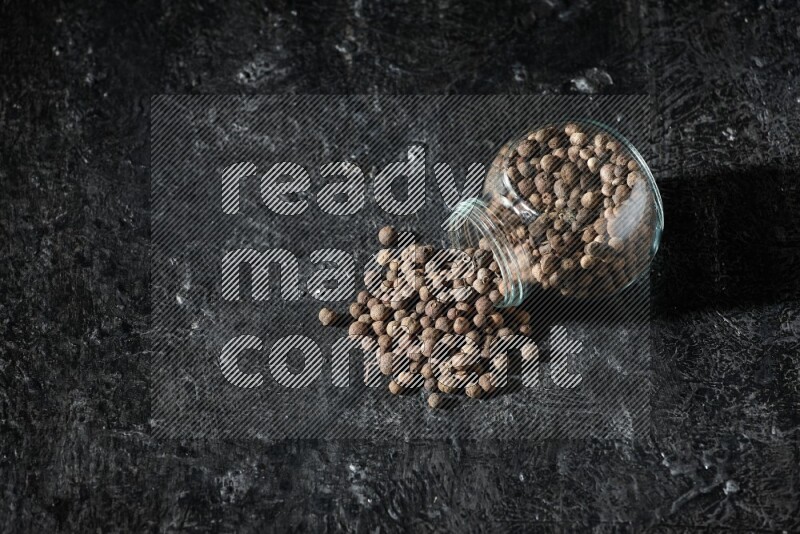 وعاء زجاجي للتوابل مقلوب ممتلئ بحبوب بهار الفلفل الحلو والحبوب تناثرت منه على خلفية سوداء