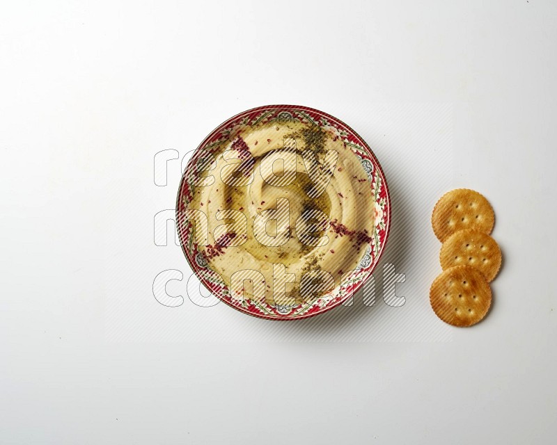 Hummus in a red plate with patterns garnished with zaatar & sumak on a white background