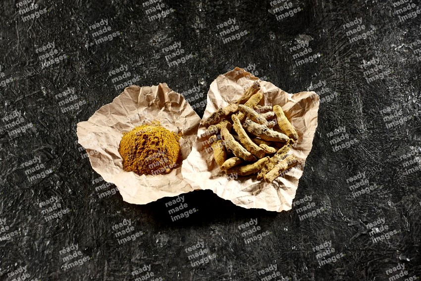 Turmeric powder and dried turmeric whole fingers in 2 crumpled pieces of craft paper on textured black flooring