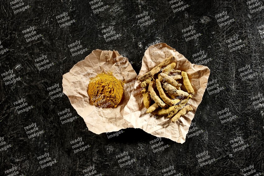 Turmeric powder and dried turmeric whole fingers in 2 crumpled pieces of craft paper on textured black flooring