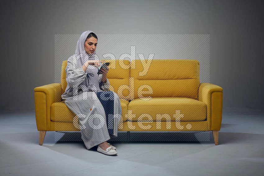 A Saudi woman wearing a light gray Abaya and white head scarf sitting on a yellow sofa and using her phone eye level on a grey background