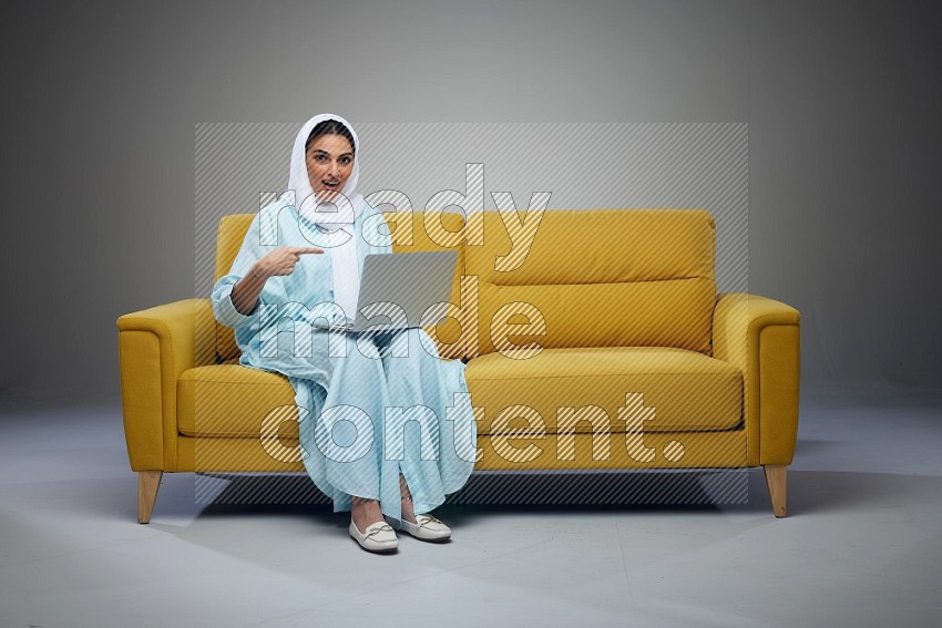 A Saudi woman sitting on a yellow Sofa With her Laptop Pointing wearing Blue Abaya